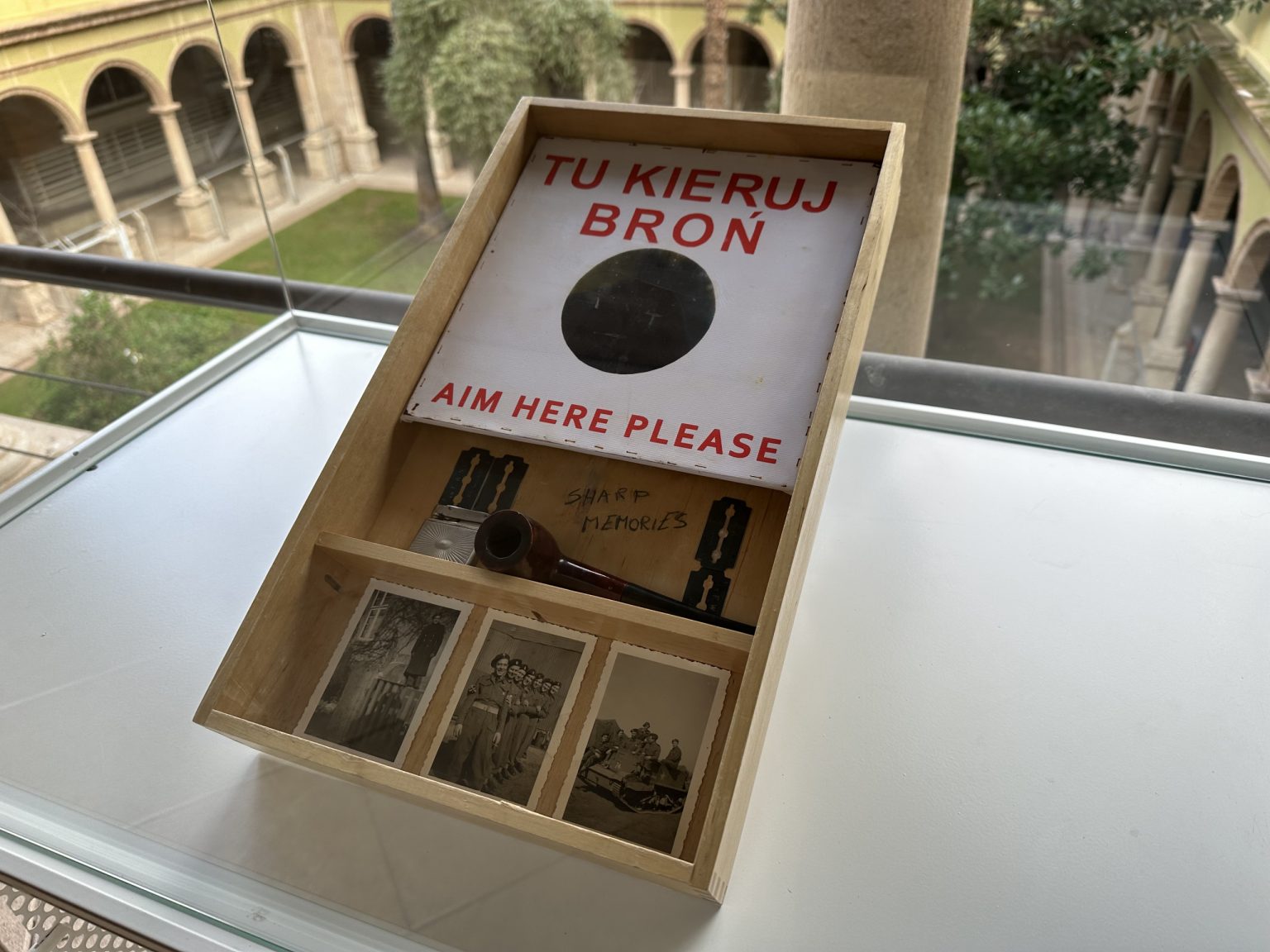 W przegródkach szuflady 3 ułożone obok siebie zdjęcia o tematyce militarnej, fajka i żyletki, oraz biały karton z napisem "Tu kieruj broń/Aim here please"