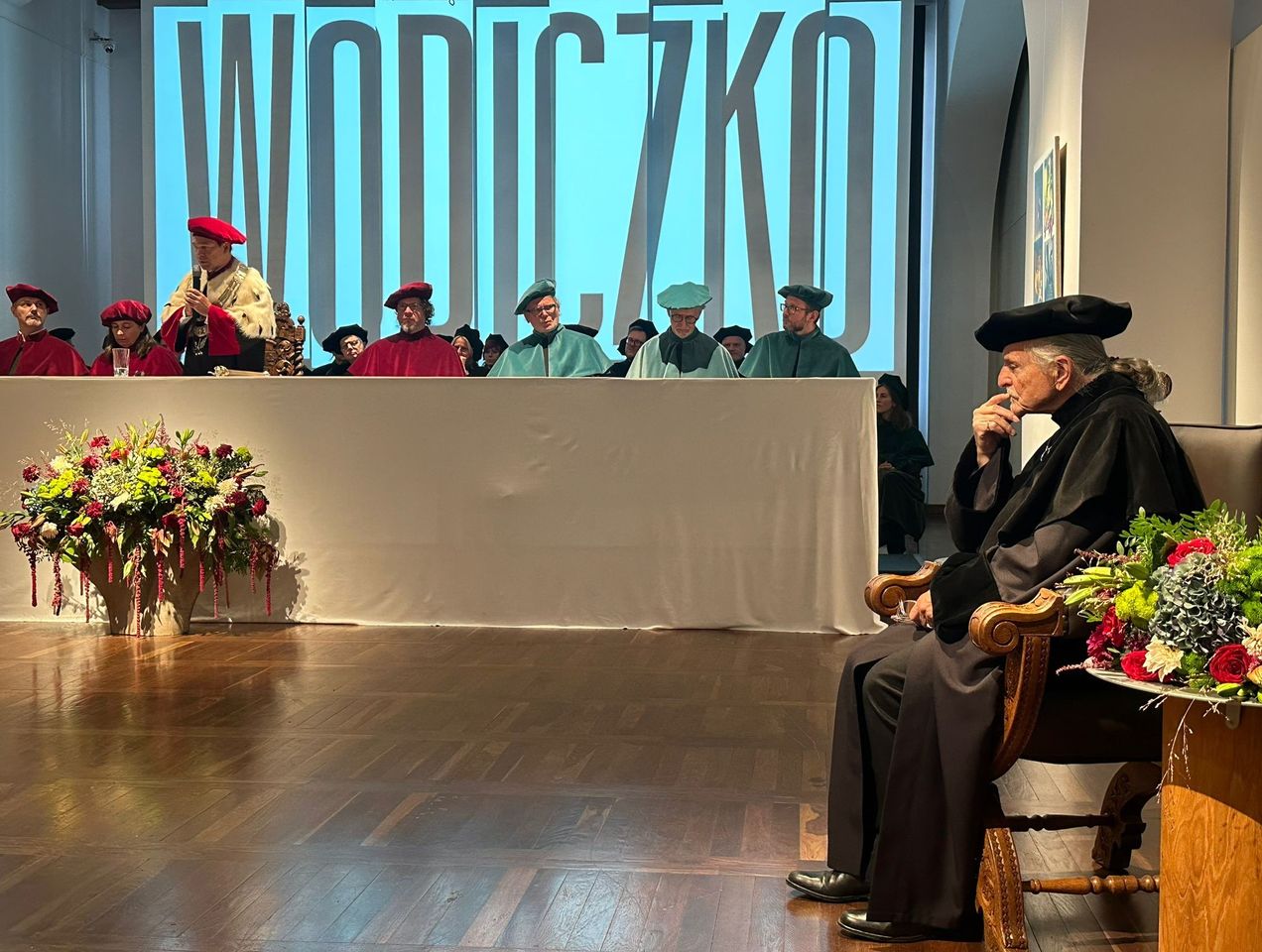 Zdjęcie z uroczystości nadania tytułu honoris causa dr Wodiczko przez Senat ASP w Gdańsku. Na zdjęciu widoczny dr Wodiczko siedzący po prawej stronie zdjęcia. W tle widać grono Senatu ASP w Gdańsku.