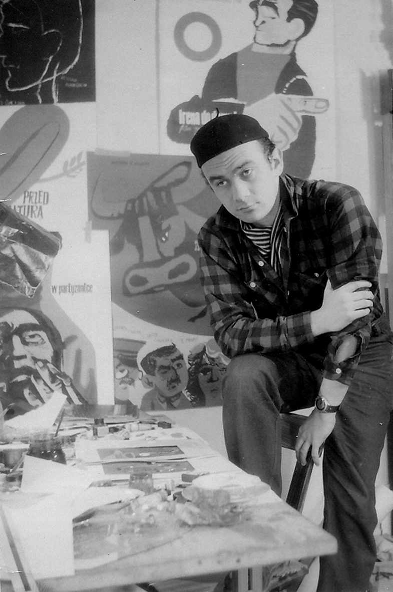 Zdjęcie Janusza Stannego z 1956r, czarnobiałe, mężczyzna w średnim wieku w czapce i kraciastej koszuli na tle swoich prac we wnętrzu pracowni