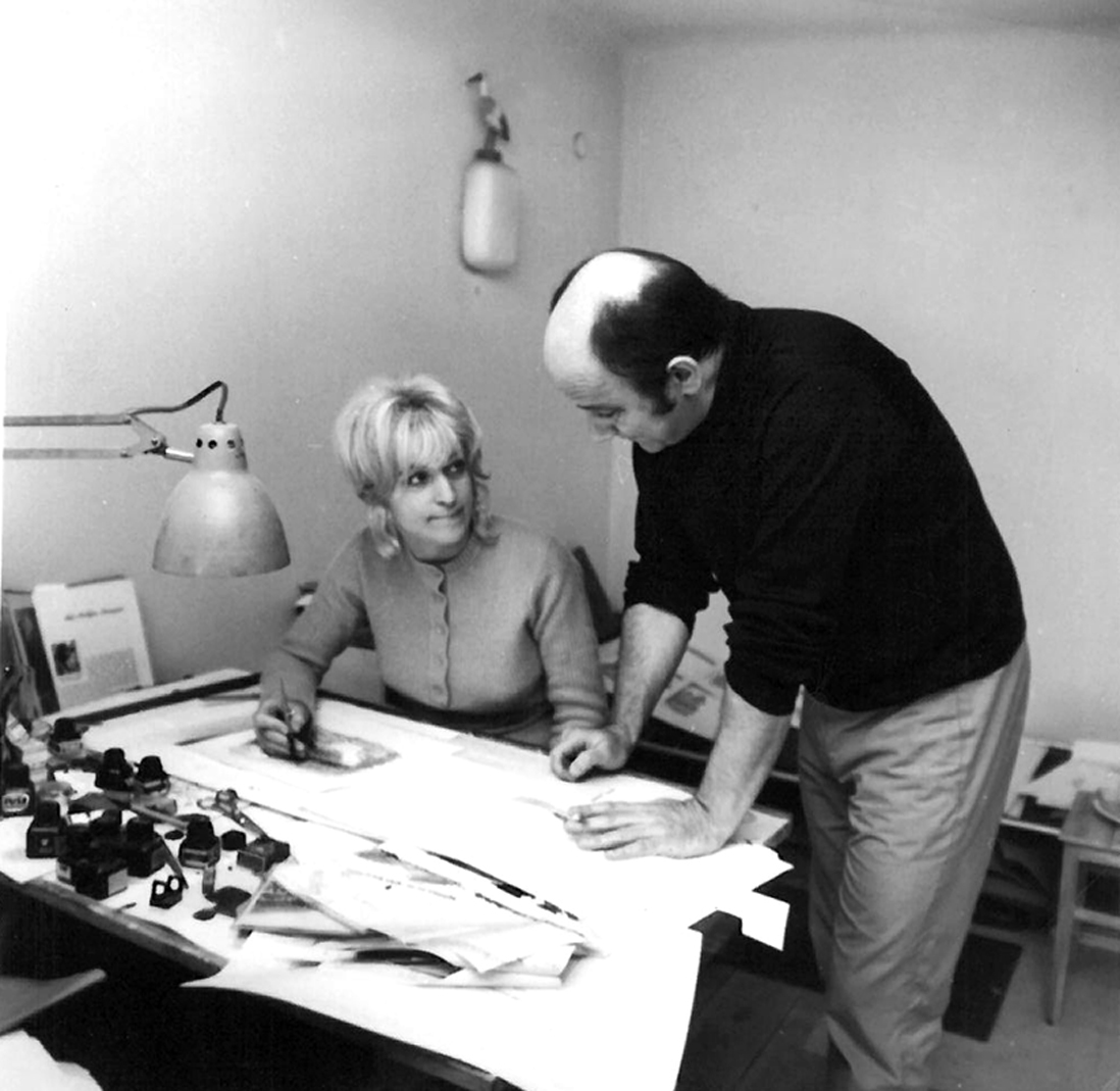 Teresa Wilbik i Janusz Stanny w pracowni w 1960 roku, fotografia czarnobiała. Teresa siedzi za biurkiem, po jej lewej stronie stoi Janusz, na biurku leżą projekty