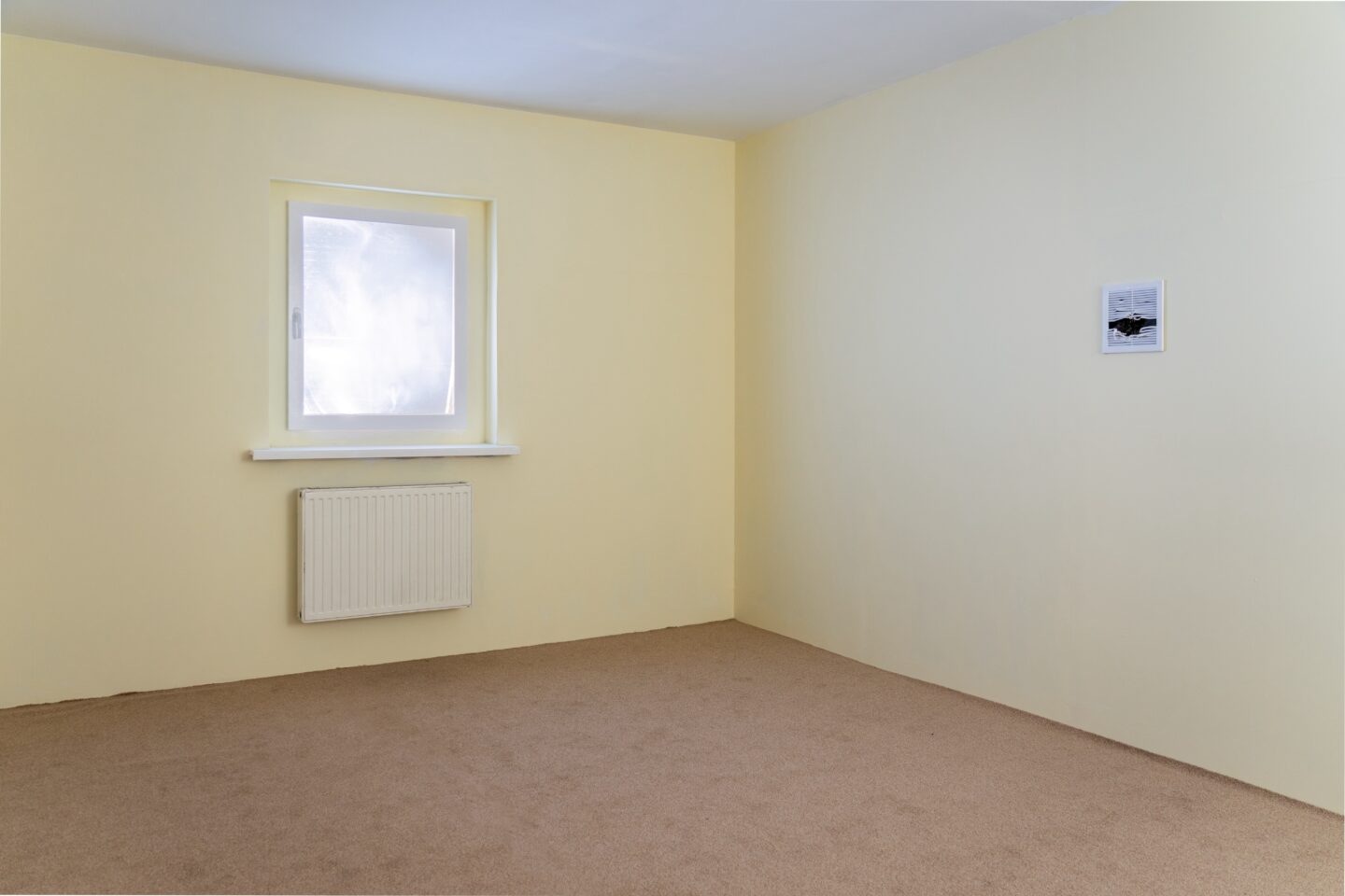 Instalacja w realistycznym rozmiarze pokoju dziennego. Pokój ma kaloryfer i okno, przez które wpada mocne światło. Na podłodze wykładzina, ściany w żółtym odcieniu.