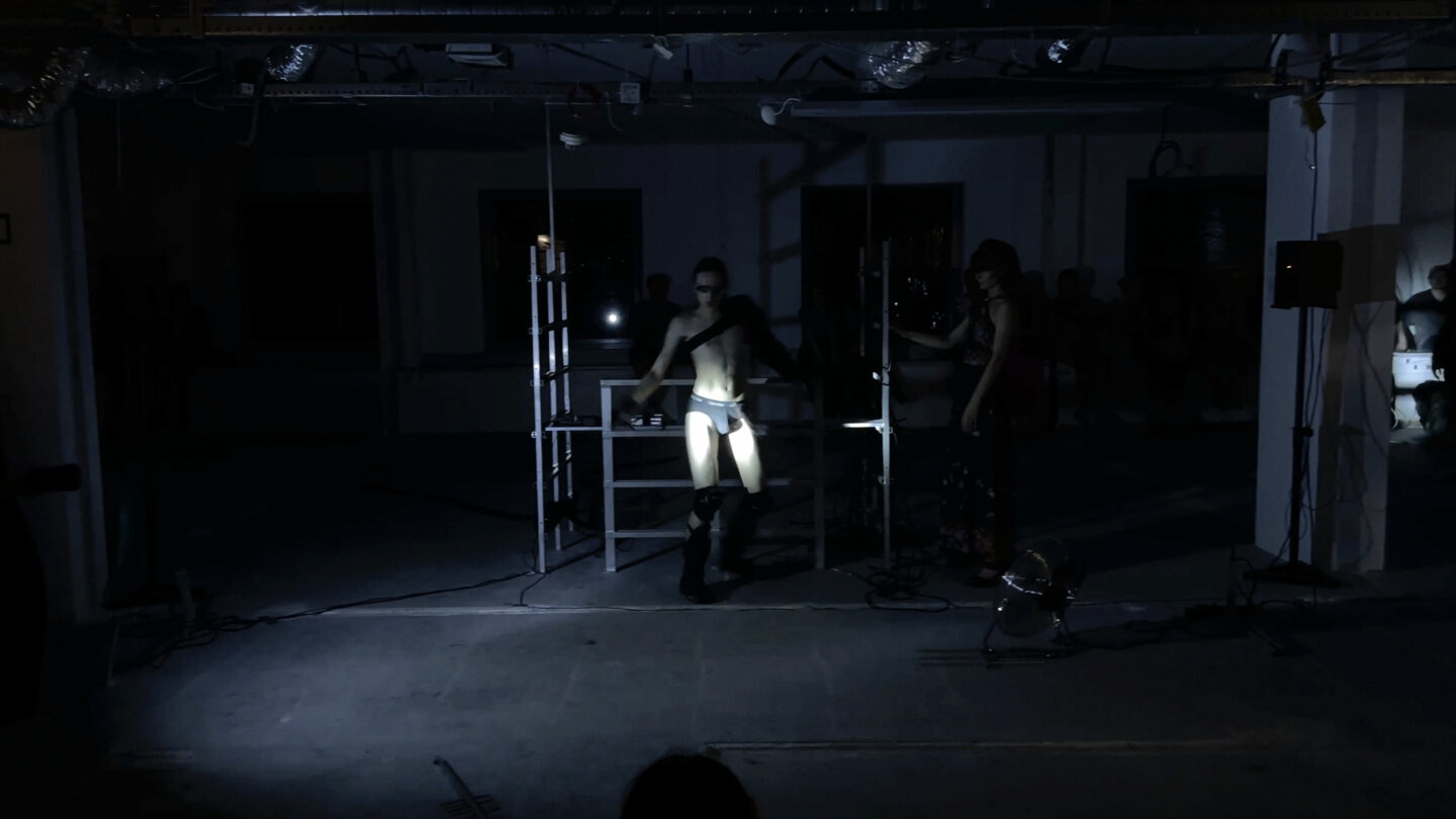 Wyciemniona przestrzeń, w tle której widać metalowe konstrukcje i oświetloną mocnym światłem postać performera.