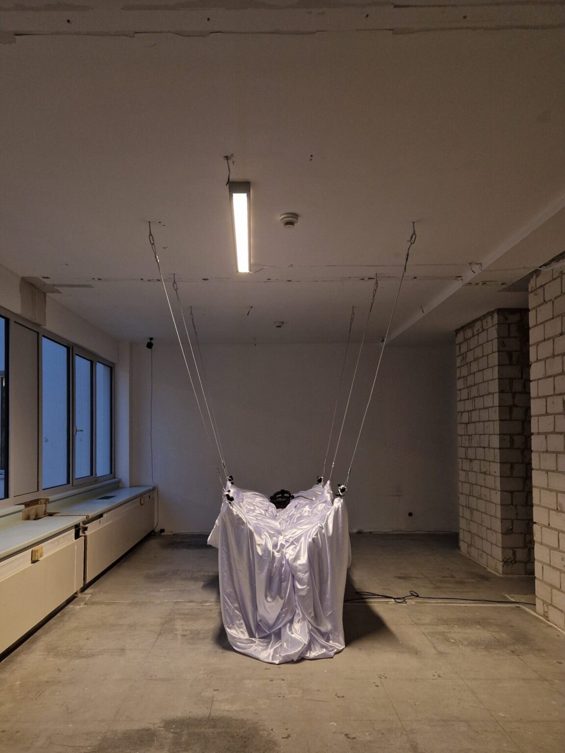 Od sufitu podwieszony obiekt przypominający łóżko, nakryty błyszczącym białym materiałem.