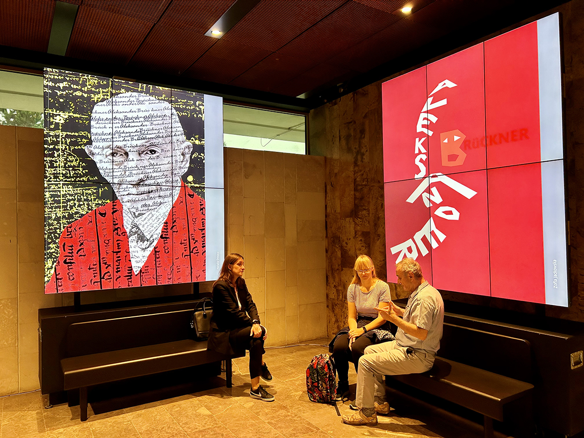 dwa plakaty z portretami prof. Aleksandra Brucknera wyświetlone na dużych ekranach w holu Biblioteki Narodowej