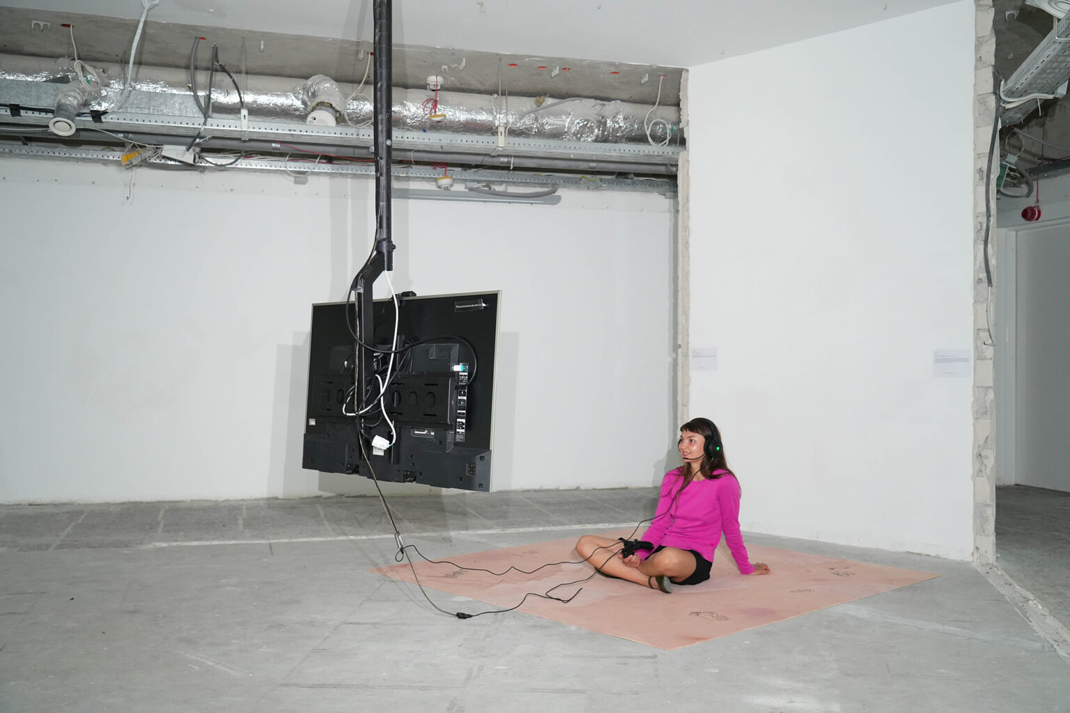 Kobieta w różowej bluzie siedzi przed telewizorem na dywanie położonym na betonowej podłodze w industrialnym pomieszczeniu. Na głowie ma słuchawki z mikrofonem.