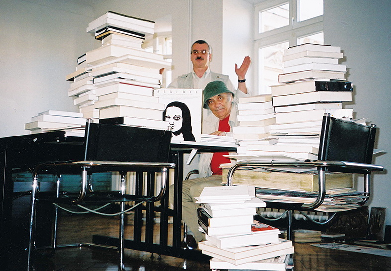 Janusz Stanny i Zygmunt Januszewski w Pracowni Ilustracji. Dwóch mężczyzn siedzi przy stole, na którym piętrzą się książki.