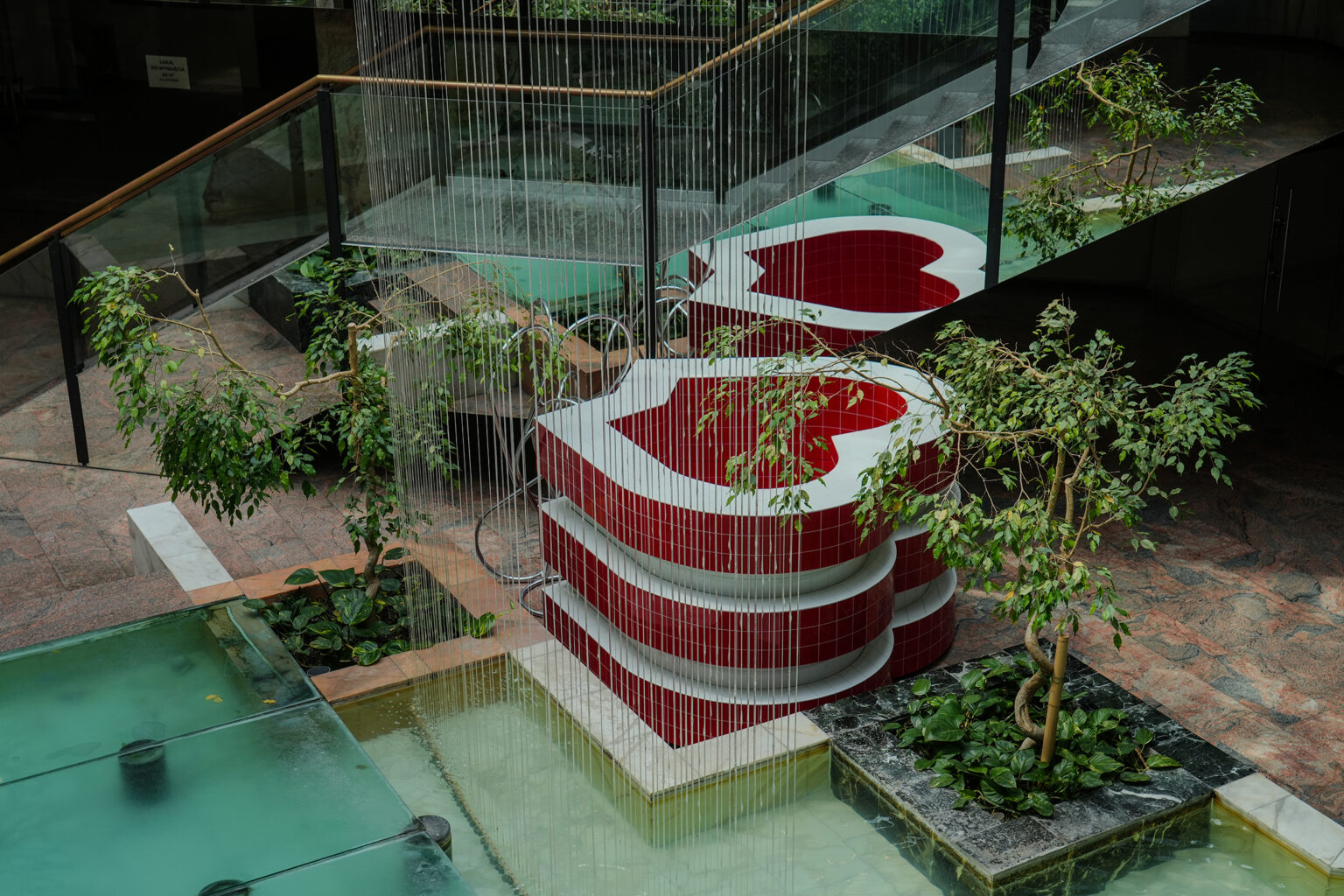 Czerwono-białe rzeźby w kształcie serc stoją przy schodach nad wodą, otoczone roślinami i strumieniami wody.