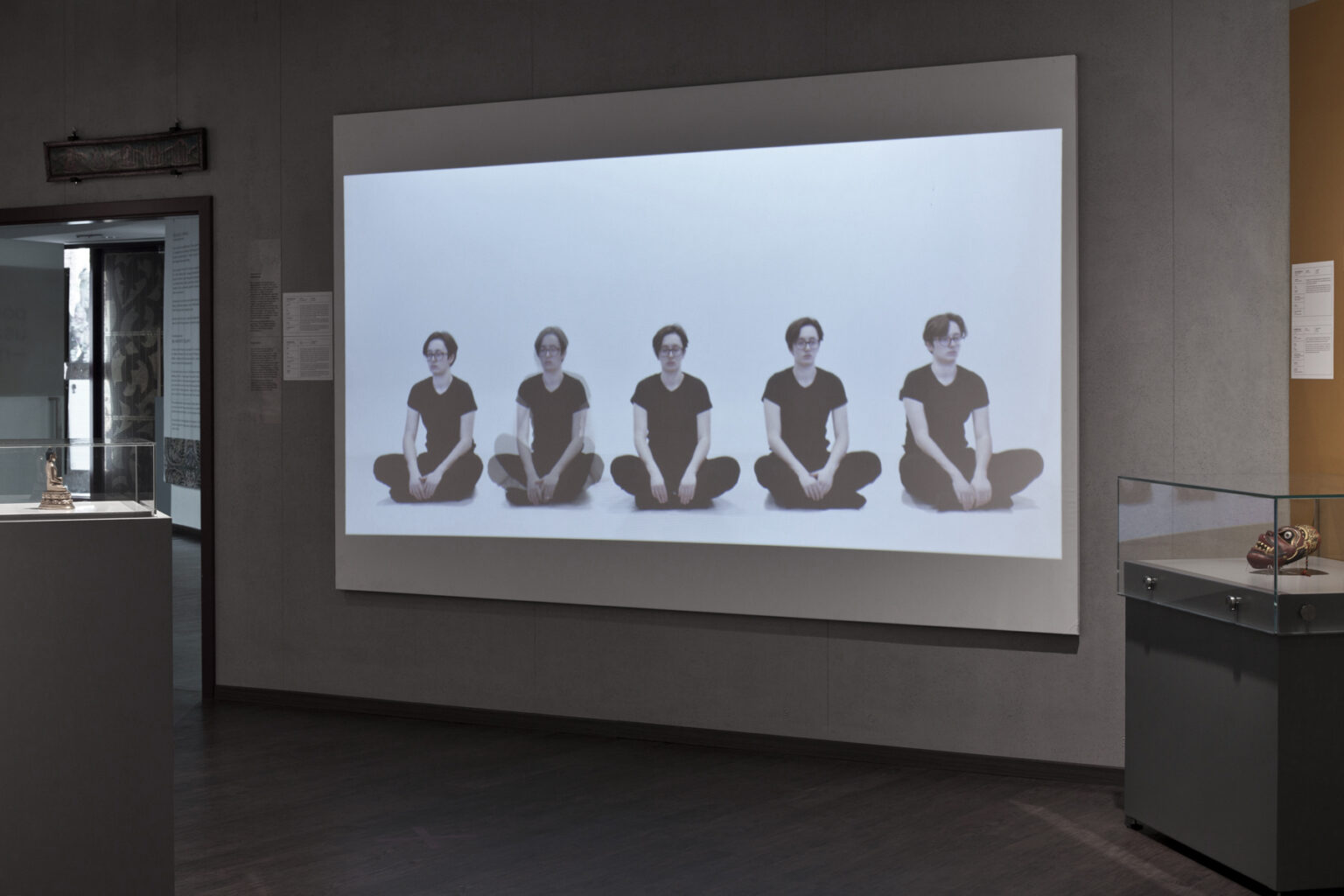 Na ścianie muzeum wyświetlony jest film z pięcioma identycznymi osobami siedzącymi po turecku na białym tle.