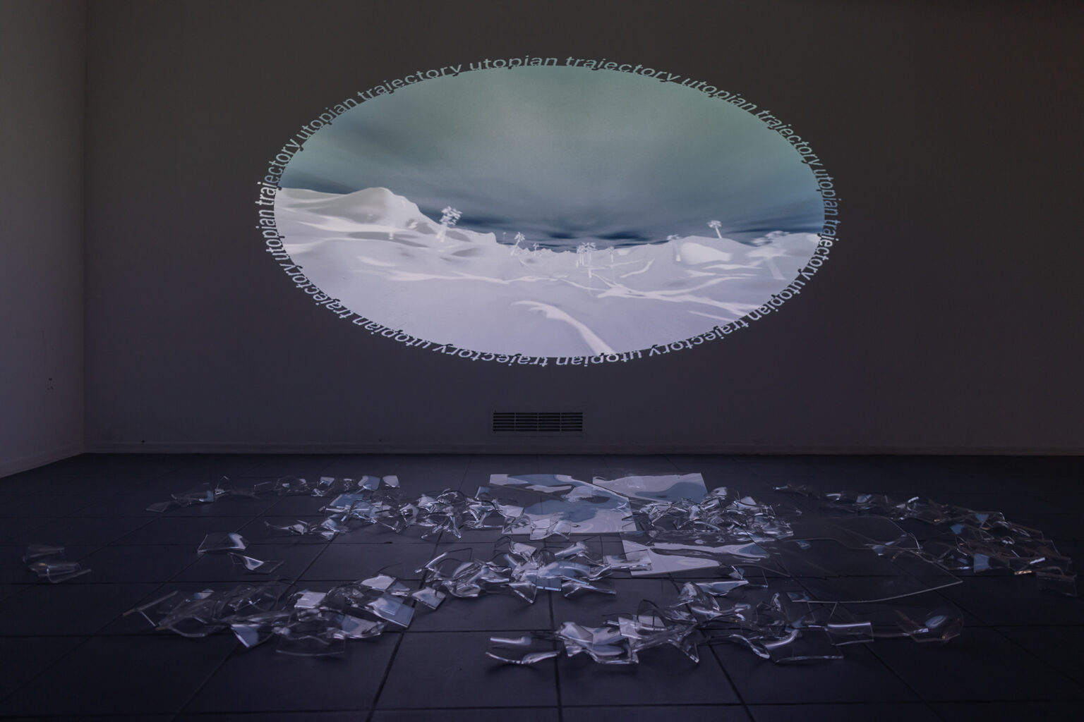 Na ścianie okrągła projekcja pejzażu z napisem „utopian trajectory”, na podłodze leżą przezroczyste fragmenty plastiku.