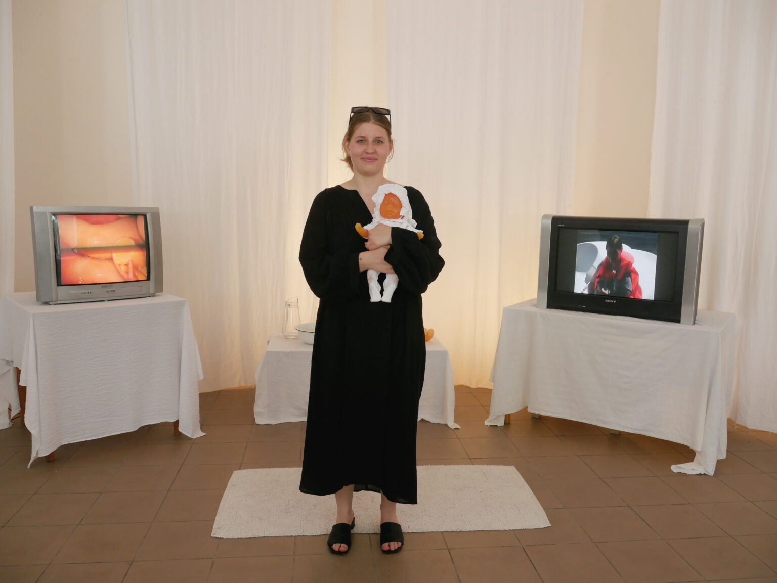 Kobieta w czarnej sukni stoi między dwoma telewizorami i trzyma lalkę owiniętą w biały materiał.