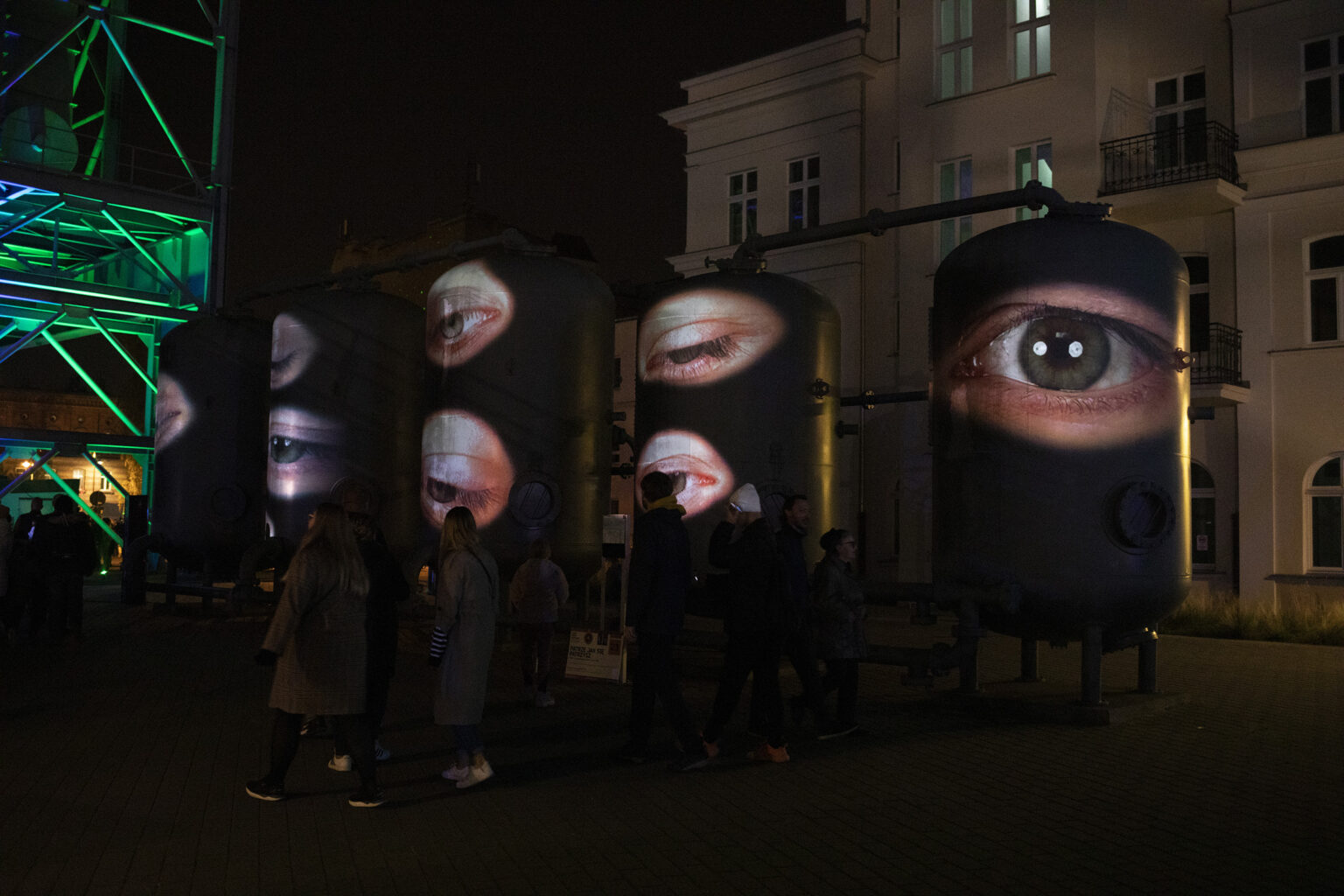 Nocna projekcja dużych oczu na cylindrycznych zbiornikach, obok przechodzą ludzie w ciemnych ubraniach.