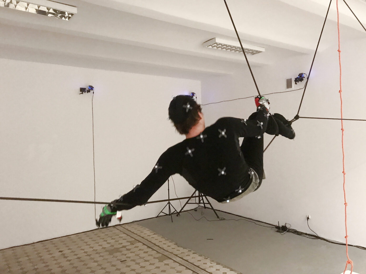 osoba w czarnym kostiumie przeznaczonym do systemu Motion Capture jest zawieszona na linach w przestrzeni sali.