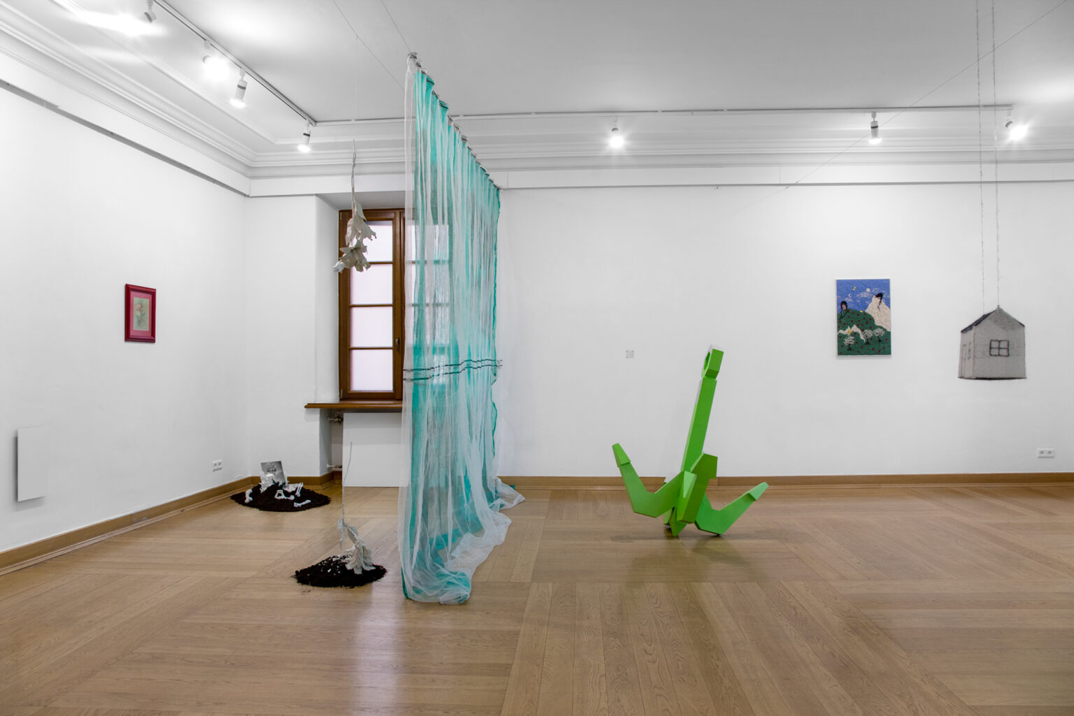 Jasna sala galerii z instalacjami: zieloną rzeźbą, błękitną tkaniną, obrazami i modelami domów.