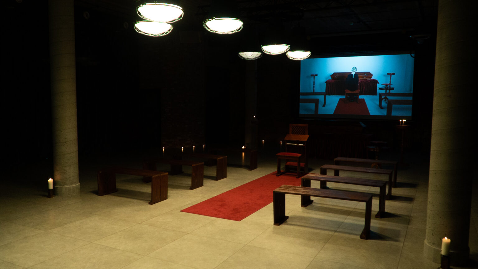 Ciemna sala z ławkami i czerwonym dywanem, na ekranie projekcja osoby siedzącej przed trumną.