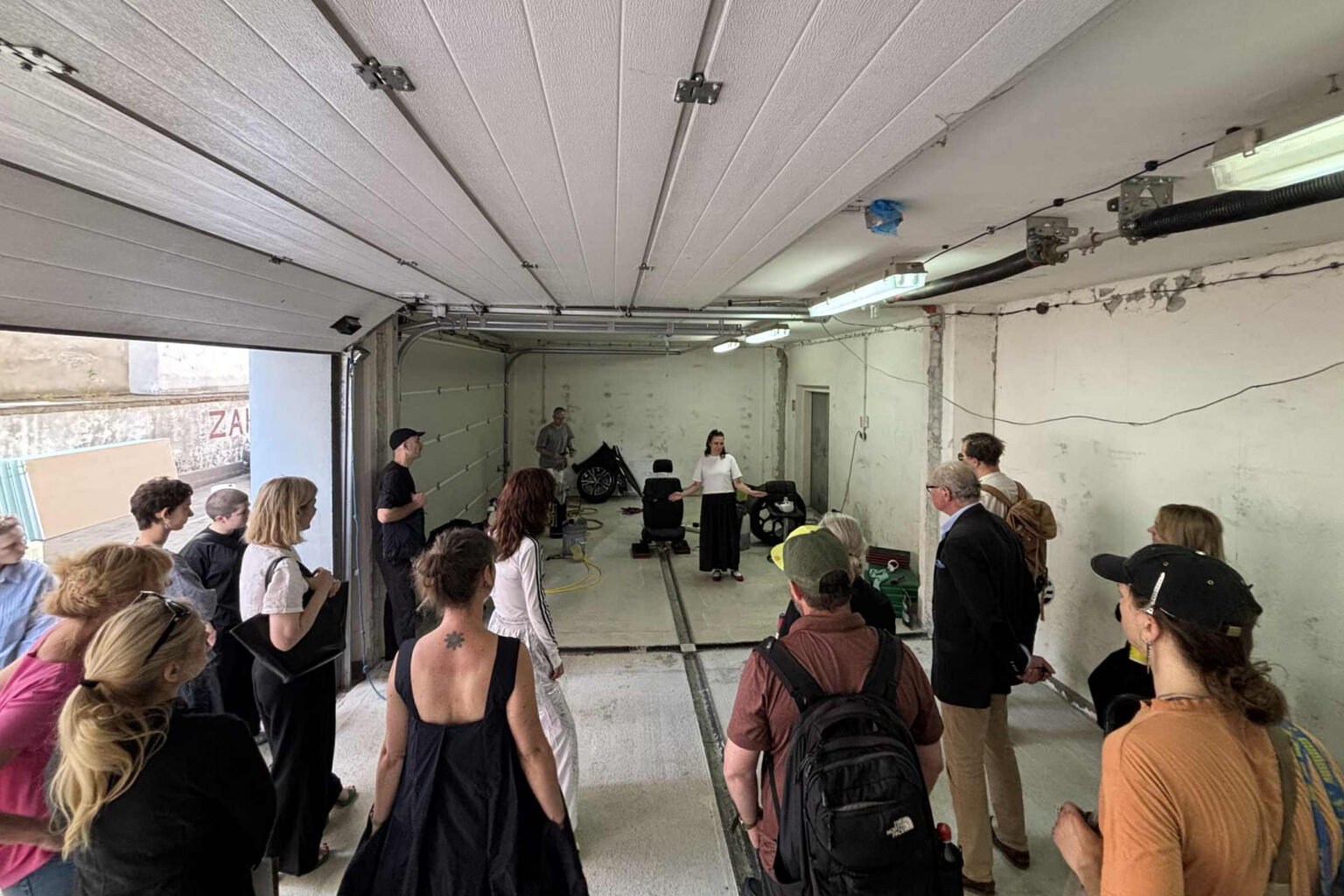 Grupa osób stoi w garażu, słuchając kobiety w białej bluzce i czarnej spódnicy, która przemawia w surowym wnętrzu.