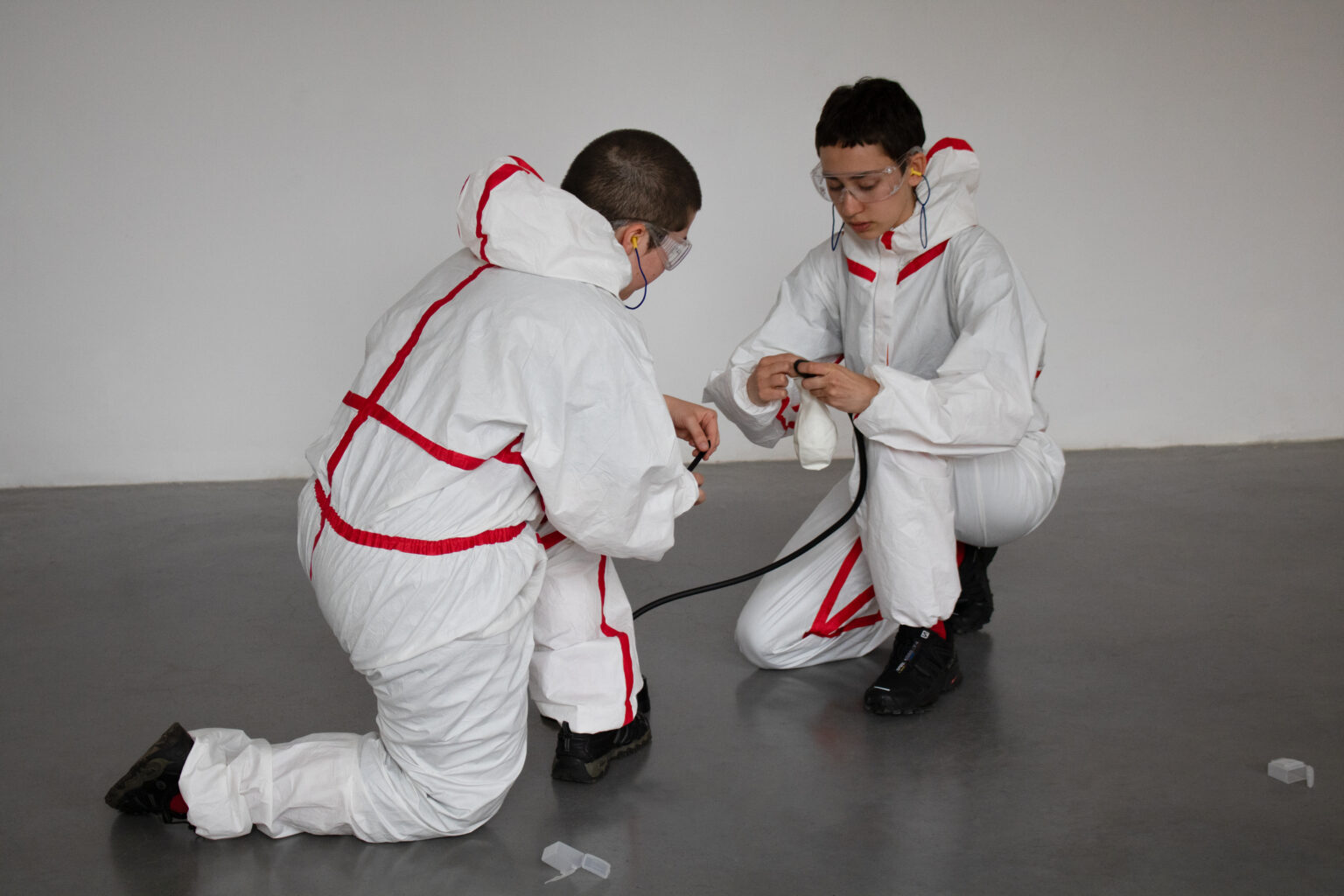 Dwie osoby w strojach ochronnych klęczą w sali wystawienniczej. Za pomocą taśmy izolacyjnej przyklejają balonik do pompki ręcznej.