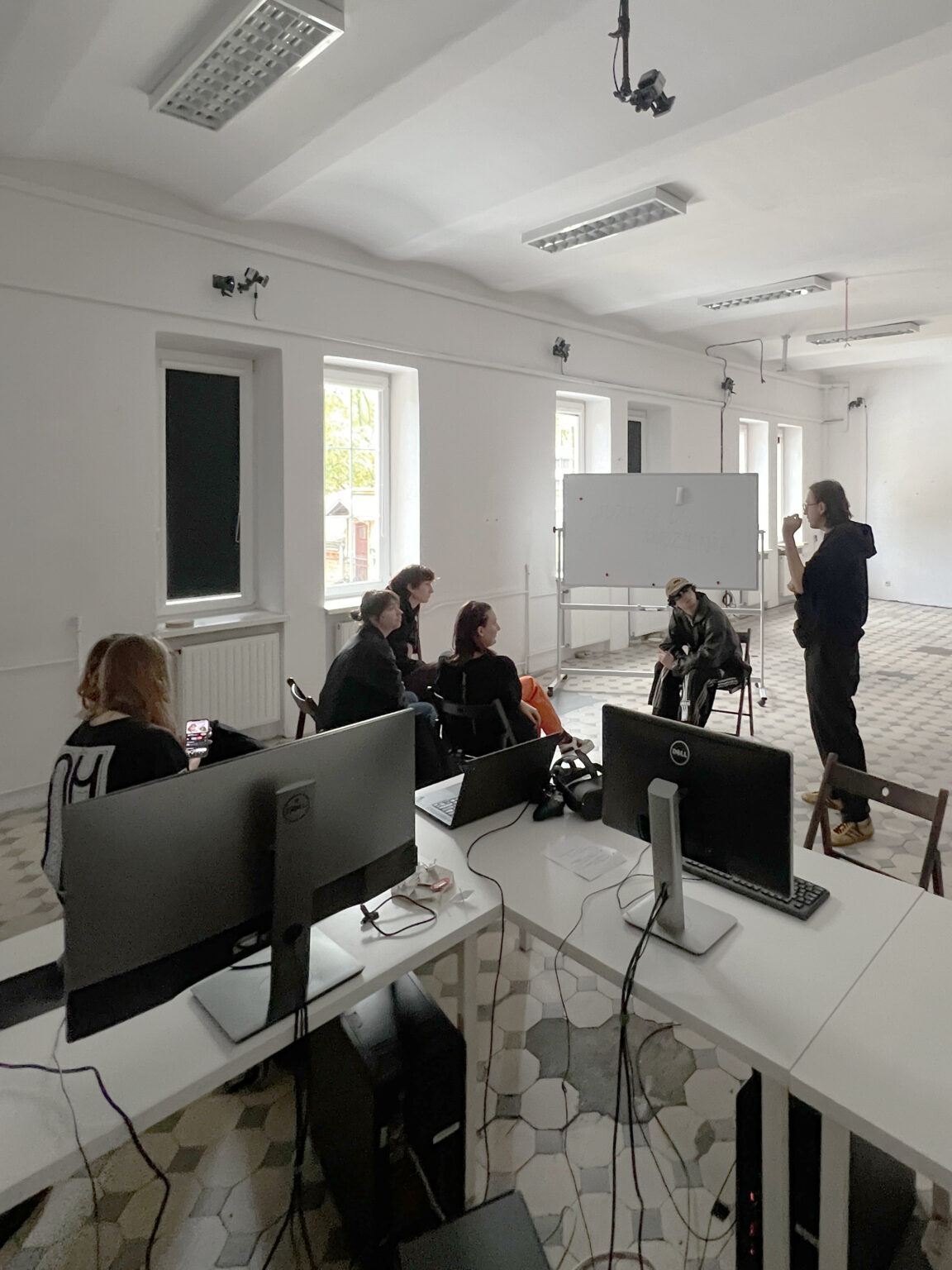 osoby siedza przy białej tablicy, jedna stoi i wykłada.