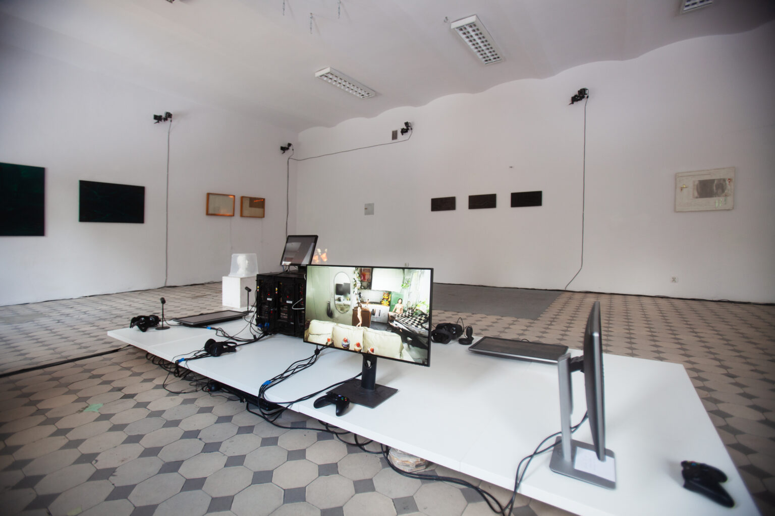 widok wystawy końcoworocznej, na pierwszym planie, telewizory, ekrany i komputery umieszczone na nisko ustawionym blacie. W tle na ścianach także prace studenckie.