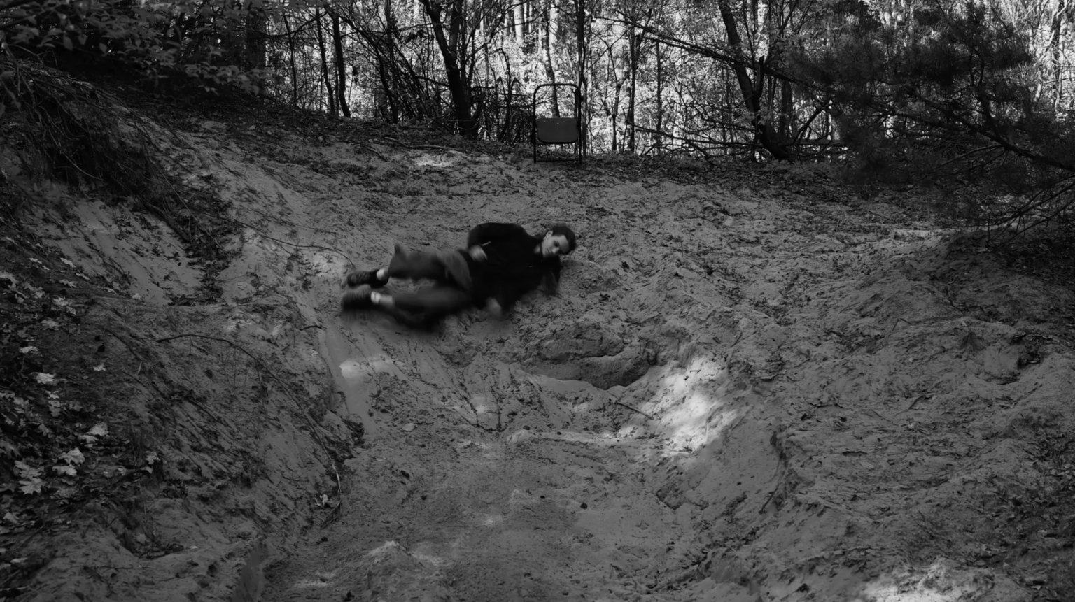 Kadr z czarno białego filmu. Osoba turla się z piaszczystego wzniesienia na szczycie, którego stoi krzesło.