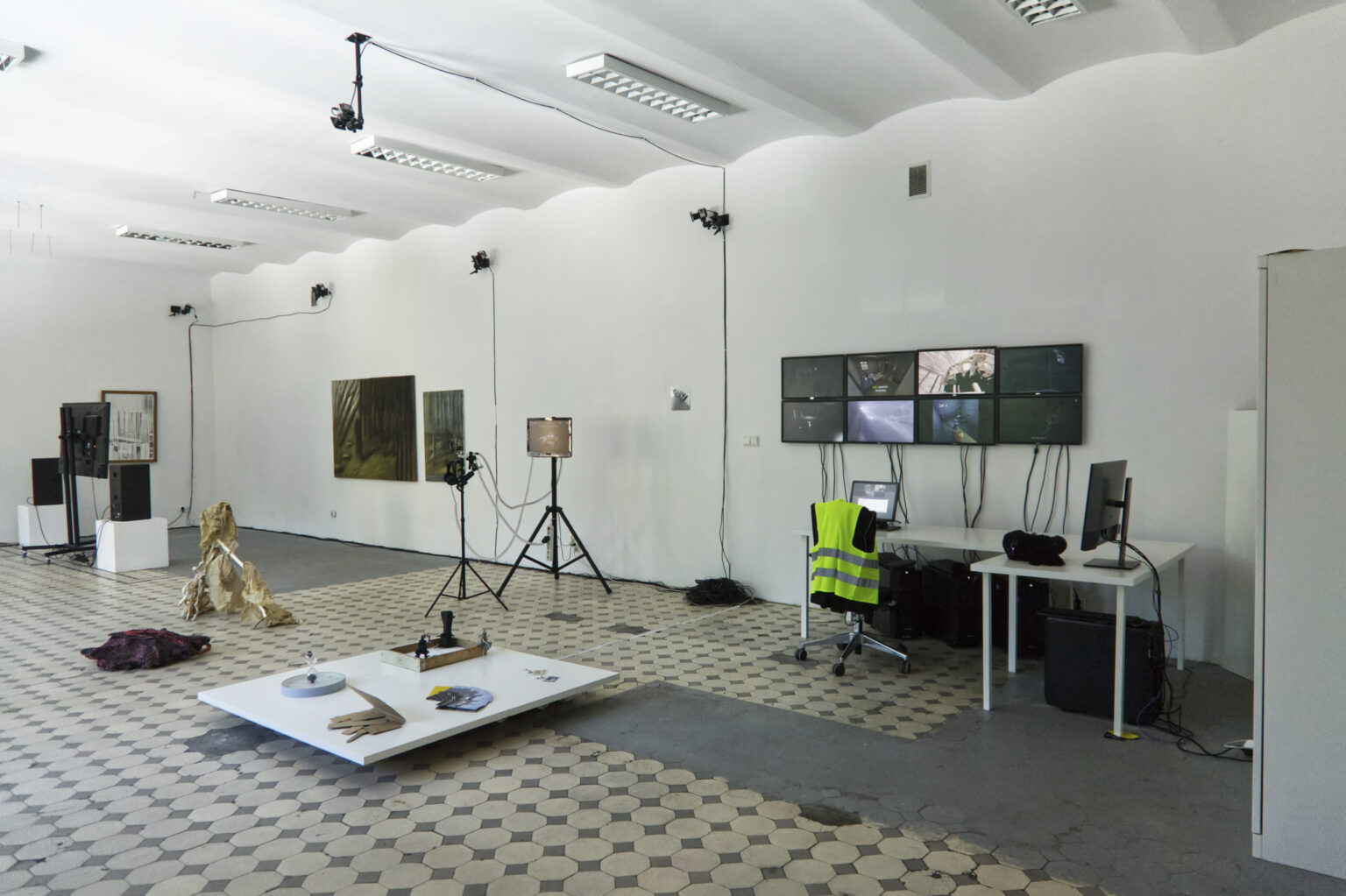 widok wystawy końcoworocznej, obiekty, ekrany na ścianie, biurko, krzesło, kamizelka ochroniarska, obrazy na ścianach