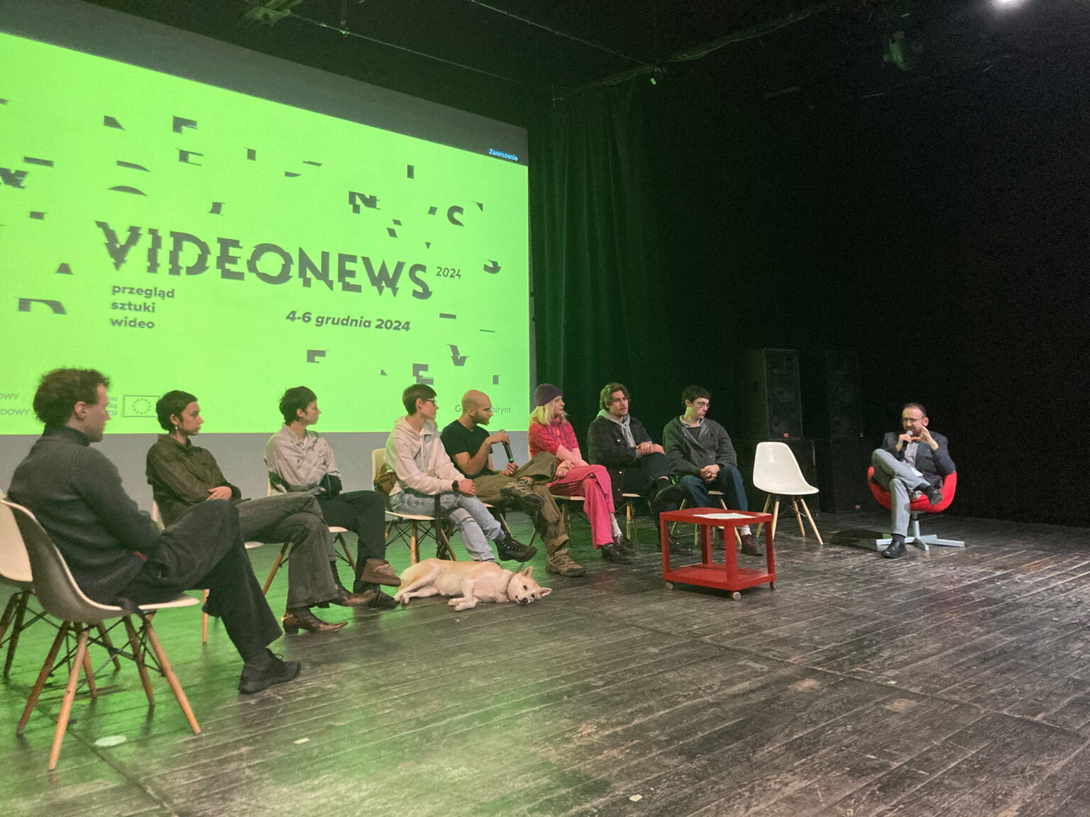 osoby siedzą przed ekranem dużej kinowej projekcji, dyskusja po projekcji prac studenckich. W tle na ekranie, duży napis VIDEONEWS, na jaskrawo-zielonym tle. Panel dyskusyjny, po proawej stronie prowadzący.