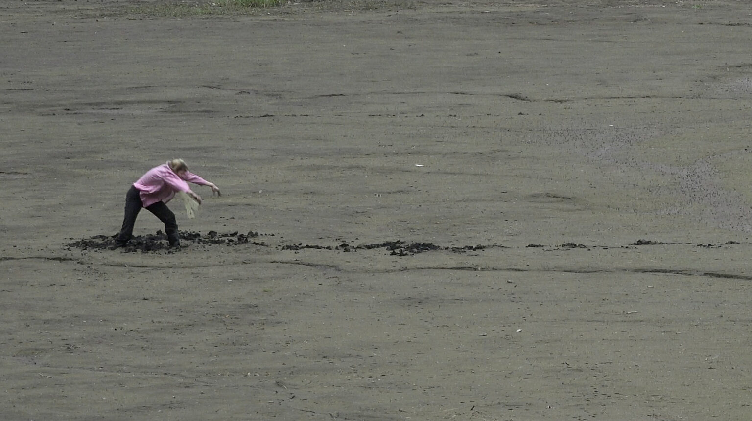 Kadr z filmu. Osoba ubrana w różową bluzkę z cekinami wykonuje ruchy taneczne stojąc po kolana w bagnie.