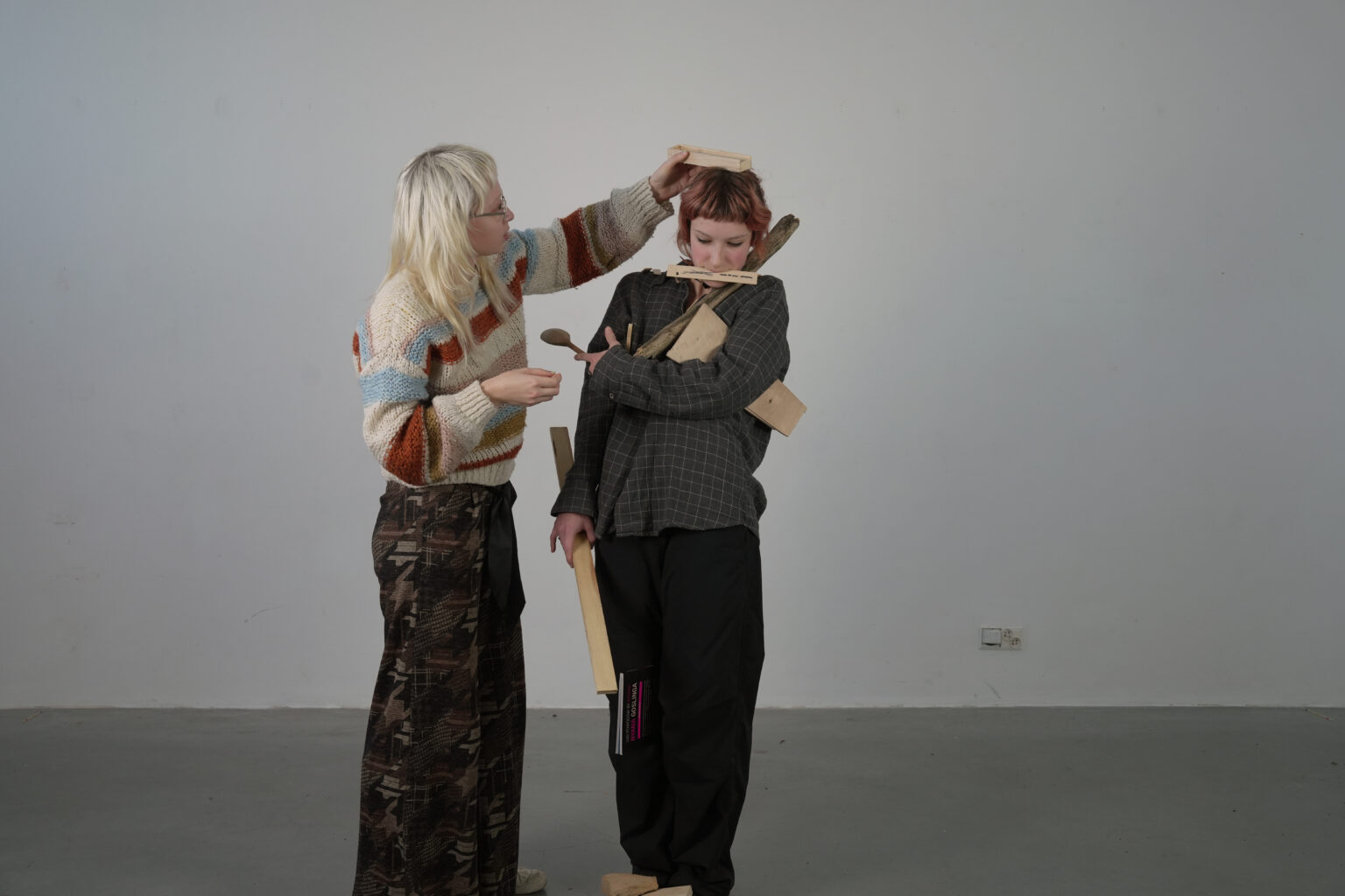 W sali wystawienniczej stoją dwie osoby. Pierwsza z nich układa kawałki drewna na głowie drugiej osoby. Druga osoba trzyma kilkanaście kawałków drewna w rękach, między rękami, między nogami, na głowie.