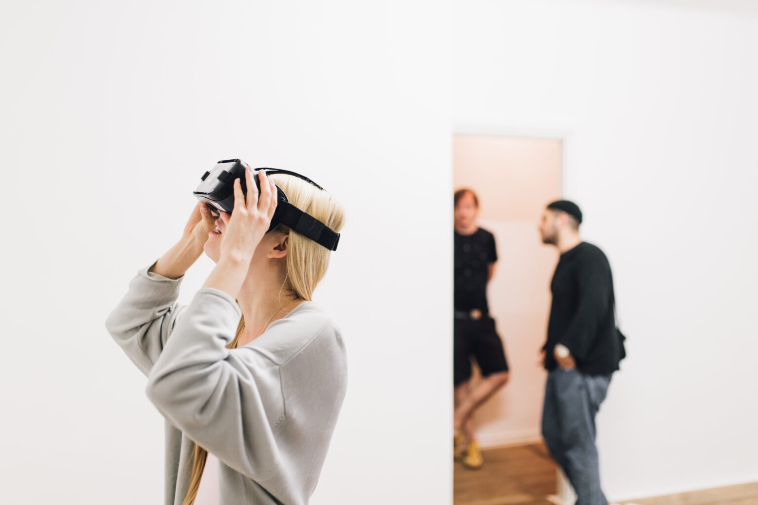 widok wystawy, osoba na pierwszym planie w goglach VR, w tle nieostro, dwie osoby rozmawiają.
