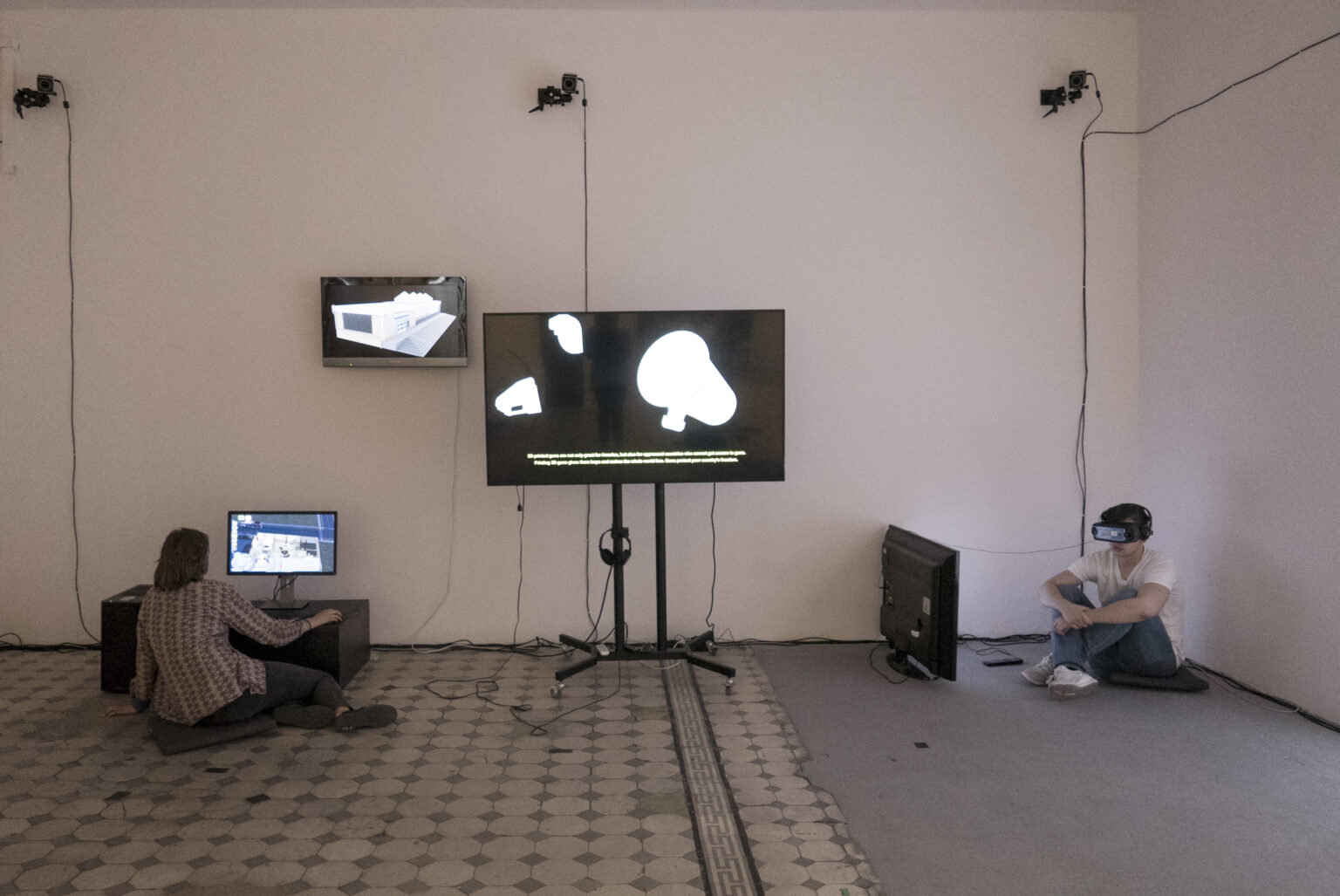 Widok wystawy końcoworocznej, wiele ekranów, na nich są pokazywane prace. Część stoi, część wisi na ścianie, dwie osoby oglądają.