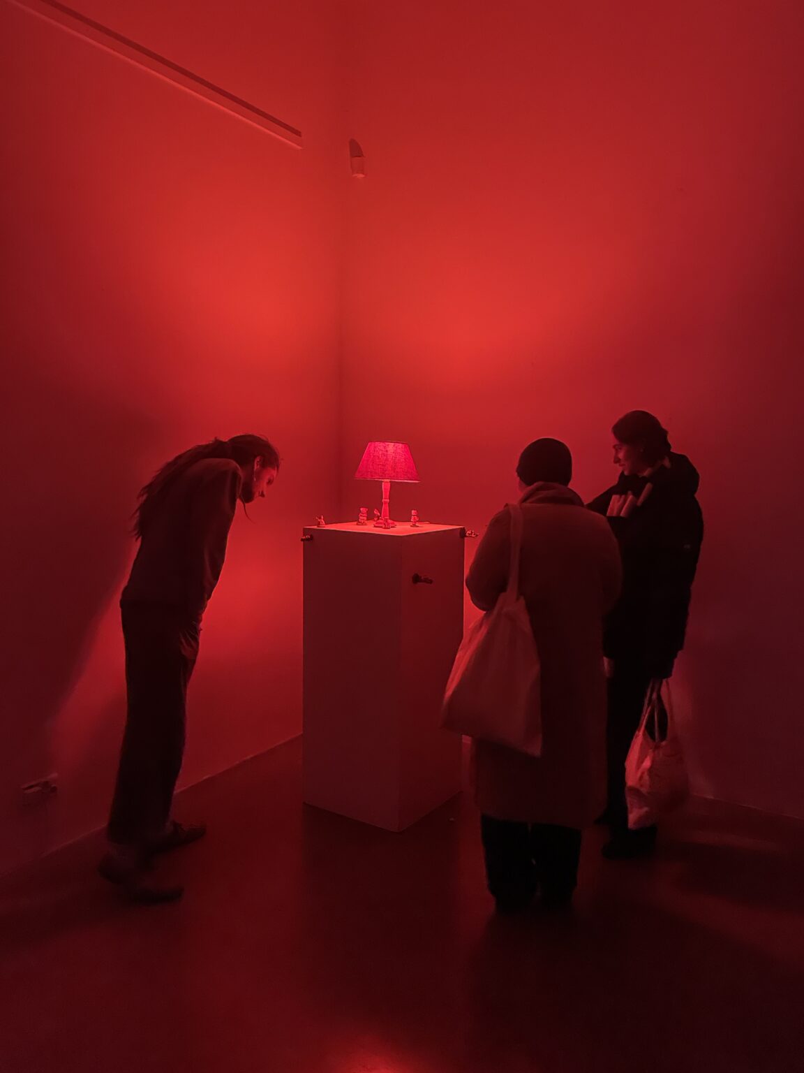 Grupa osób przygląda się małym rzeźbom stojącym na postumencie. Na postumencie stoi również czerwona lampa, która oświetla całe pomieszczenie i postacie na czerwono.