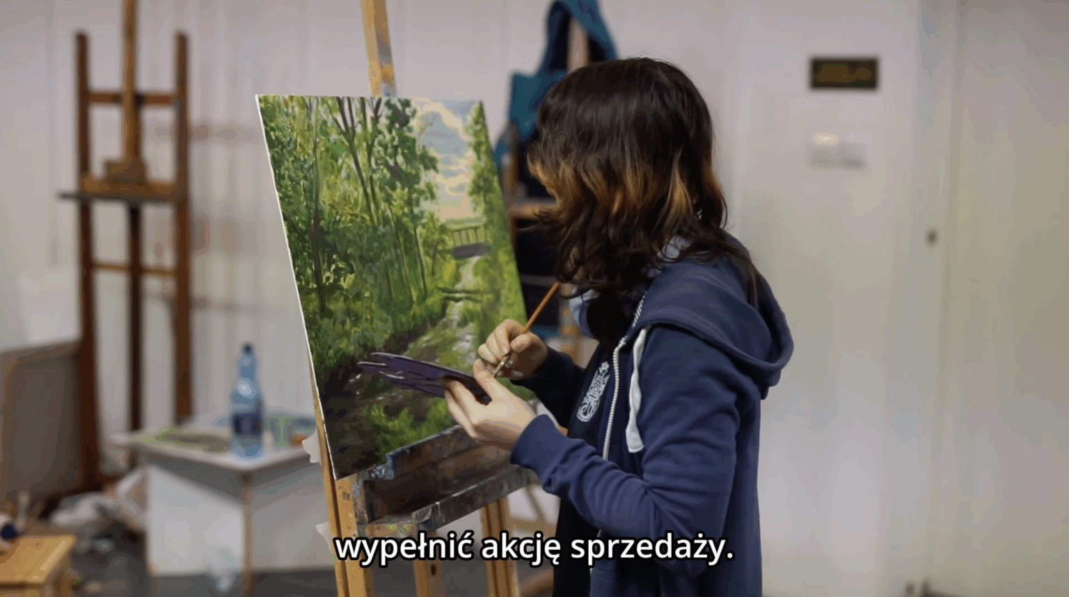 Kadr z filmu. Osoba stoi przy sztaludze i maluje obraz przedstawiający pejzaż. Na kadrze widnieje napis “wypełnić akcję sprzedaży”.