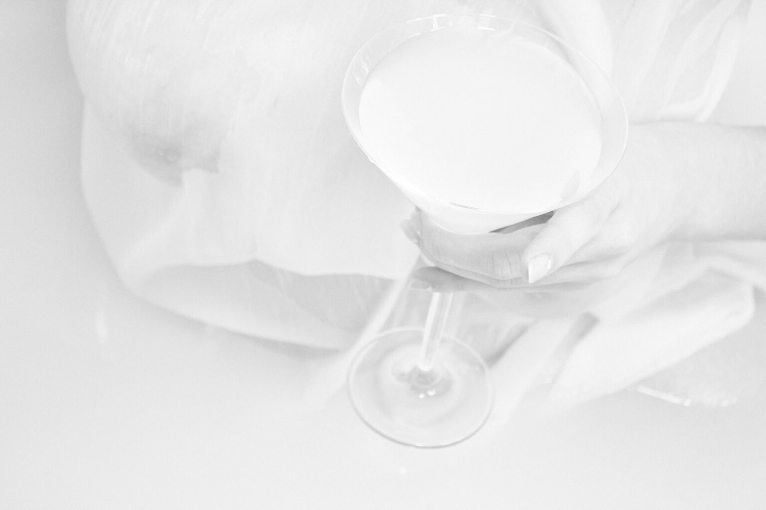ręka trzymająca kieliszek wypełniony zabieloną cieczą. W tle widać fragment nieostrej sylwetki.