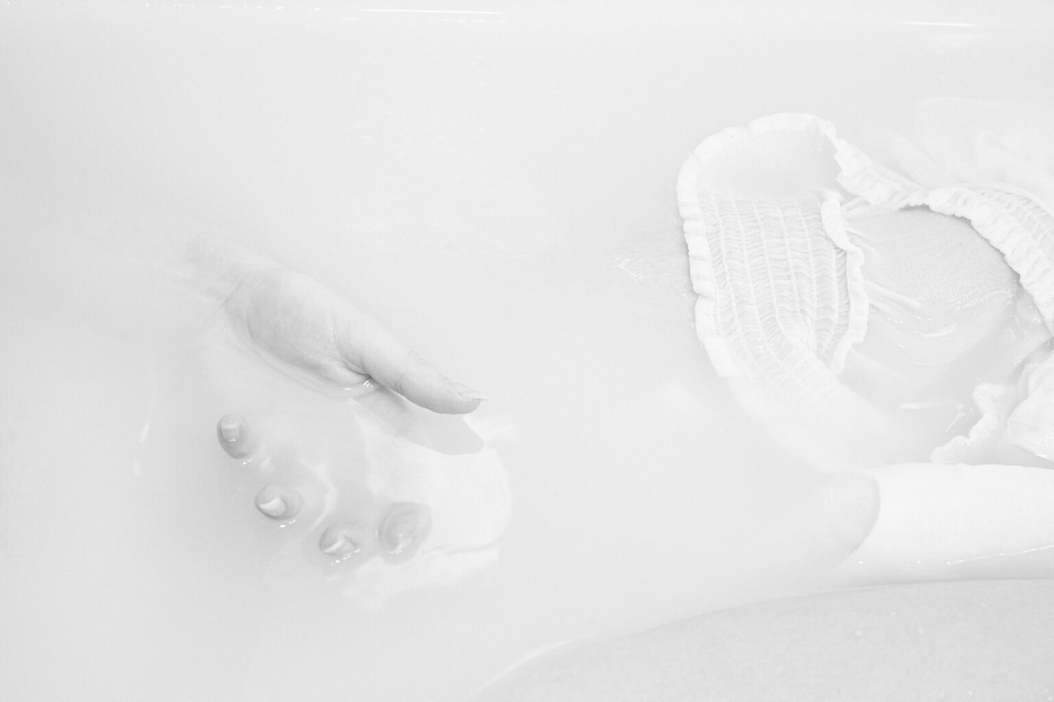 fragment ręki wynurzającej się z zabielonej cieczy. Obok widać fragment białego materiału unoszącego się na powierzchni: