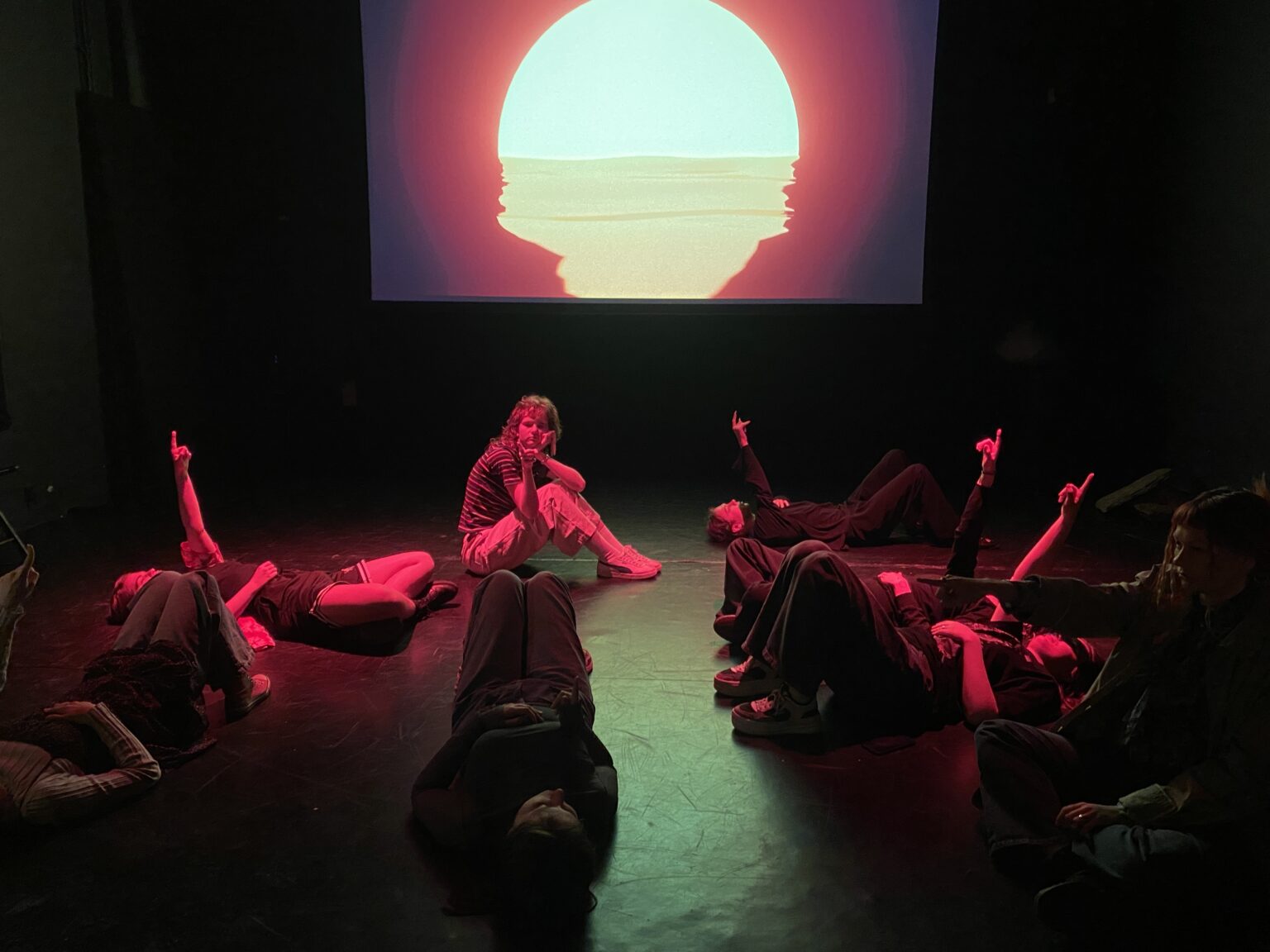Na scenie widać osoby leżące na plecach i wskazujące prawą ręką na sufit. Z tyłu sceny w tle widoczna jest projekcja zachodzącego słońca.