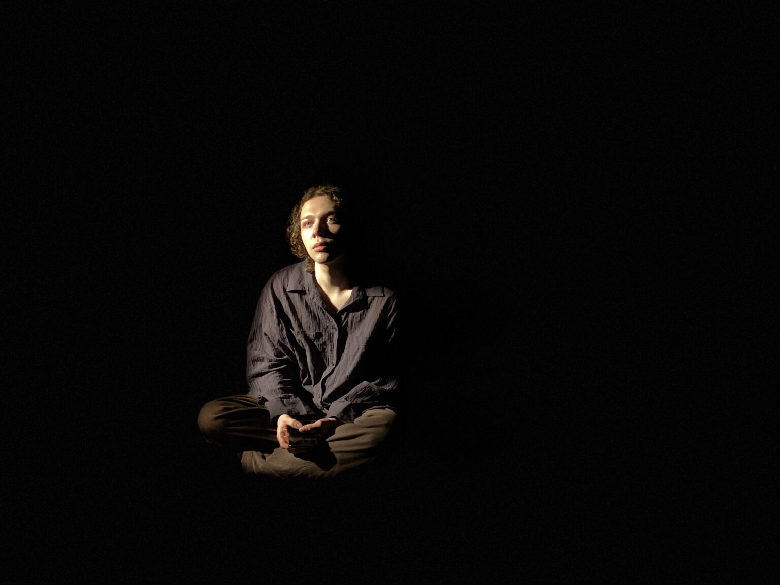 Na środku sceny z ciemności wyłania się chłopak siedzący w siadzie skrzyżnym. Jest ubrany w ciemną lnianą koszulę i ciemne spodnie. Jest oświetlony słabym ciepłym światłem kontrowobocznym z lewej strony.