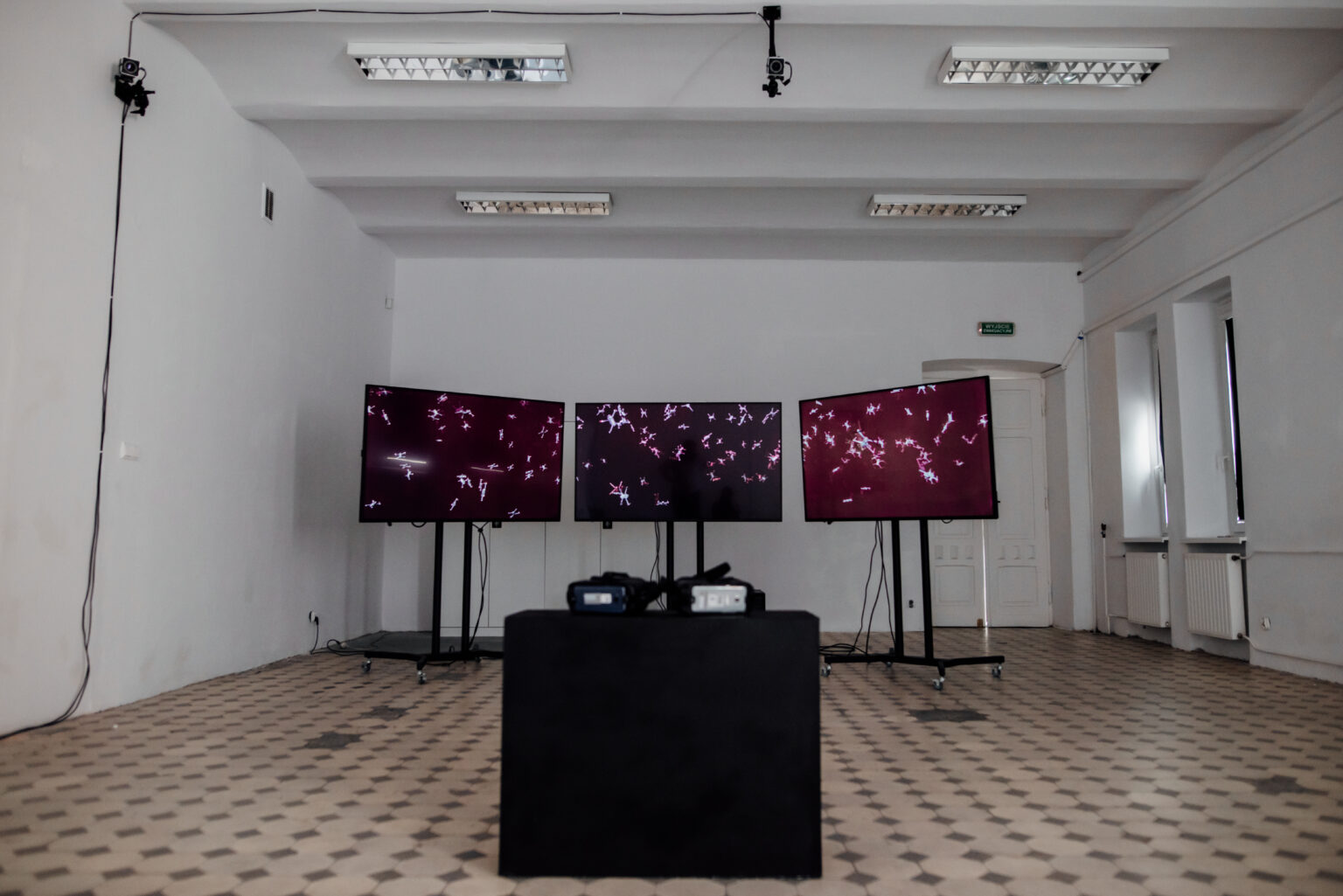 na pierwszym planie nieostro, para gogli do VR, na drugim planie 3 ekrany, na których w ciemno-burgundowych barwach wyświetlane są animacje 3D, z dużą ilością małych dronów