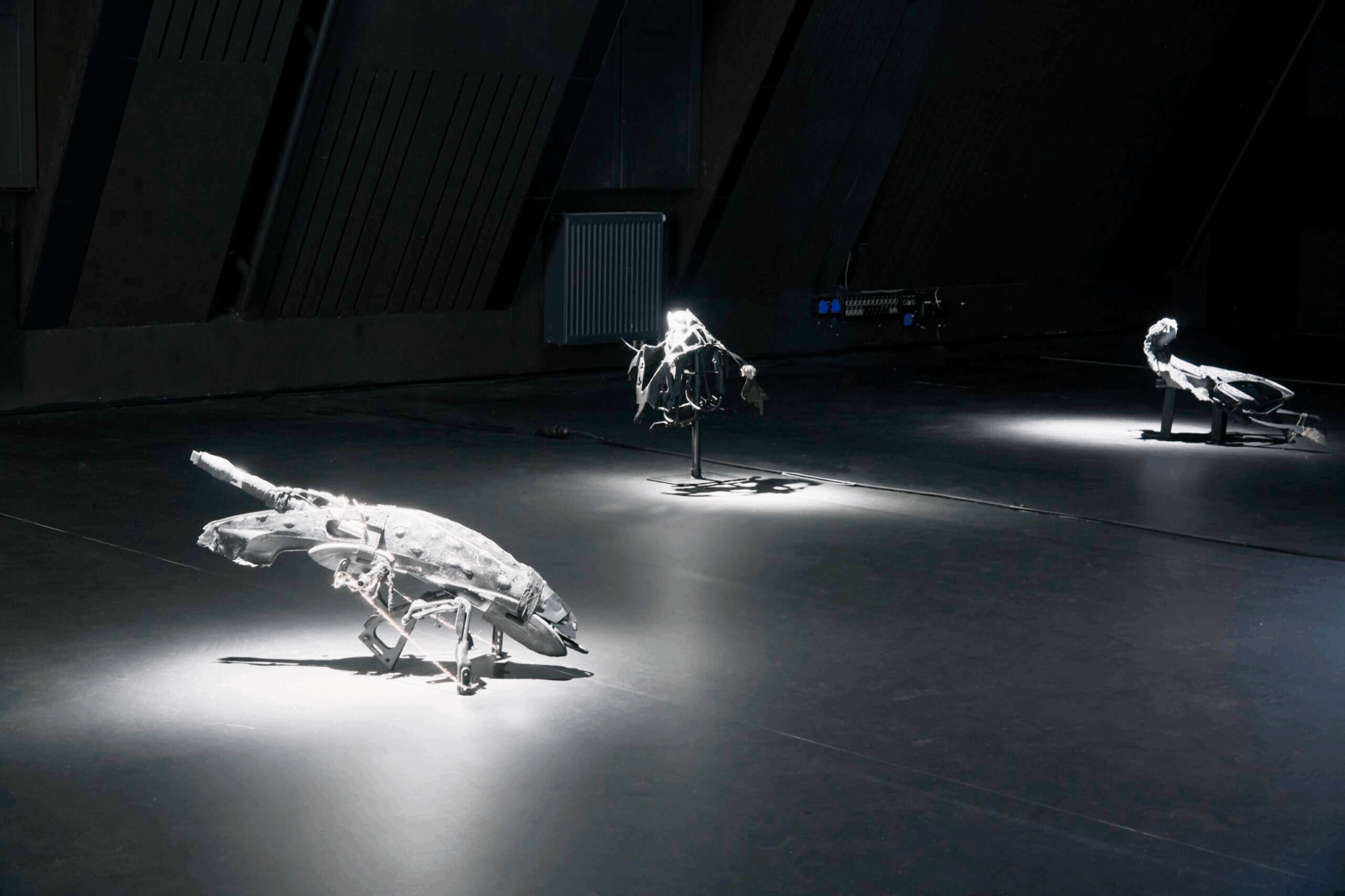 widok wystawy, mocne punktowe oświetlenie obiektów, lekko przepalone biele. Ciemnoszare i czarne obiekty na podłodze, przypominają futurystyczne, dystopijne roboty, biologiczno-mechaniczne hybrydy albo pojazdy.