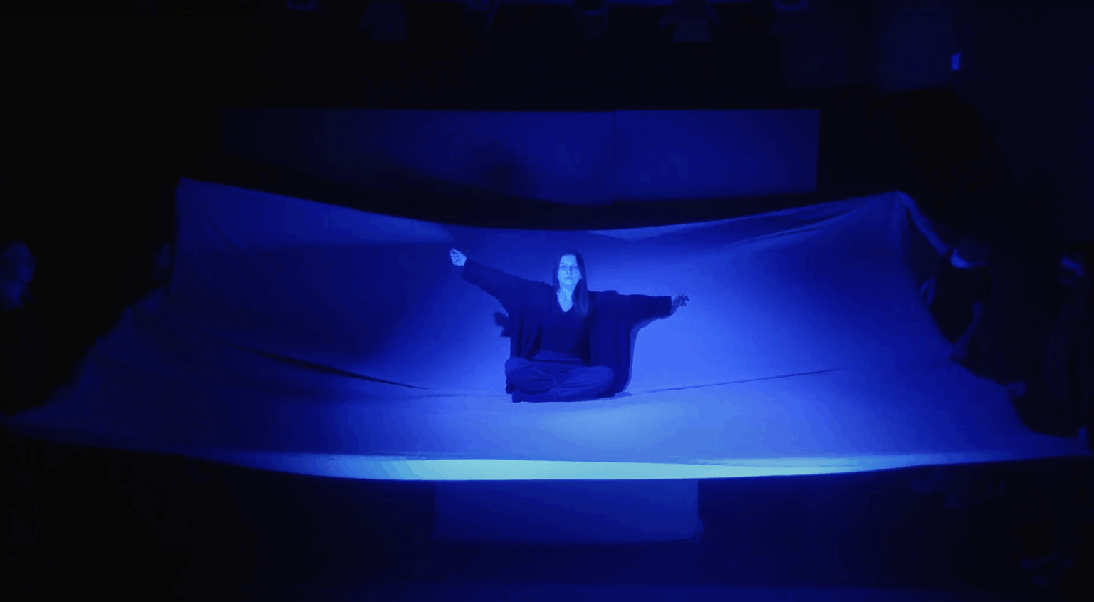 Na scenie widać wyłaniającą się z ciemności tkaninę trzymaną za brzegi przez kilka osób. Na środku tkaniny siedzi w siadzie skrzyżnym młoda dziewczyna. Pod tkaniną jest ukryty stół w sposób niewidoczny dla widza, tak żeby siedząca na tkaninie postać była stabilna. Dziewczyna ma szeroko rozłożone ramiona i wpatrzona jest przed siebie. Tkanina oraz postać oświetlone są niebieskim światłem.