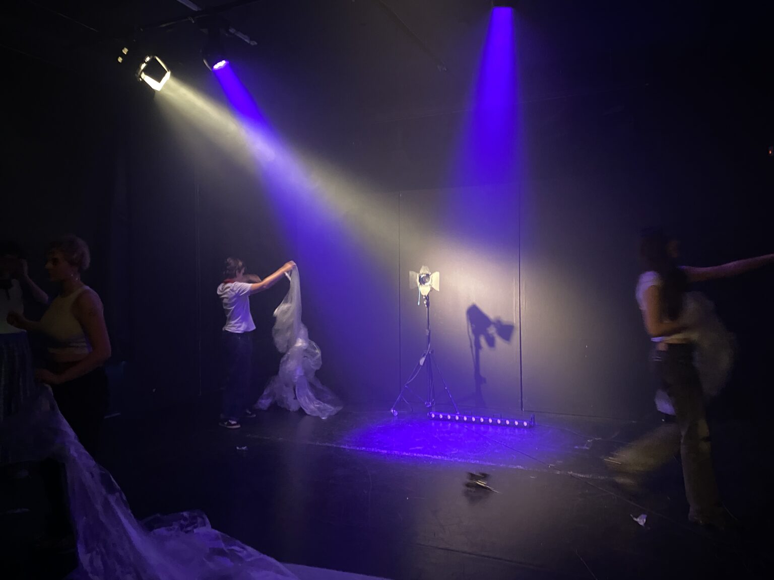 Na scenie widać trzy osoby w ruchu rozkładające folie. Na środku sceny stoi reflektor na statywie oraz ledbar na podłodze. Scena oświetlona jest snopami światła w kolorach niebieskich i żółtych.