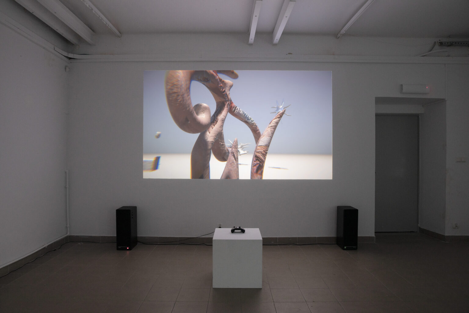 Widok instalacji, projekcja na ścianie, abstrakcyjna scena 3D, pad do gry na kubiku przed, głośniki obok.