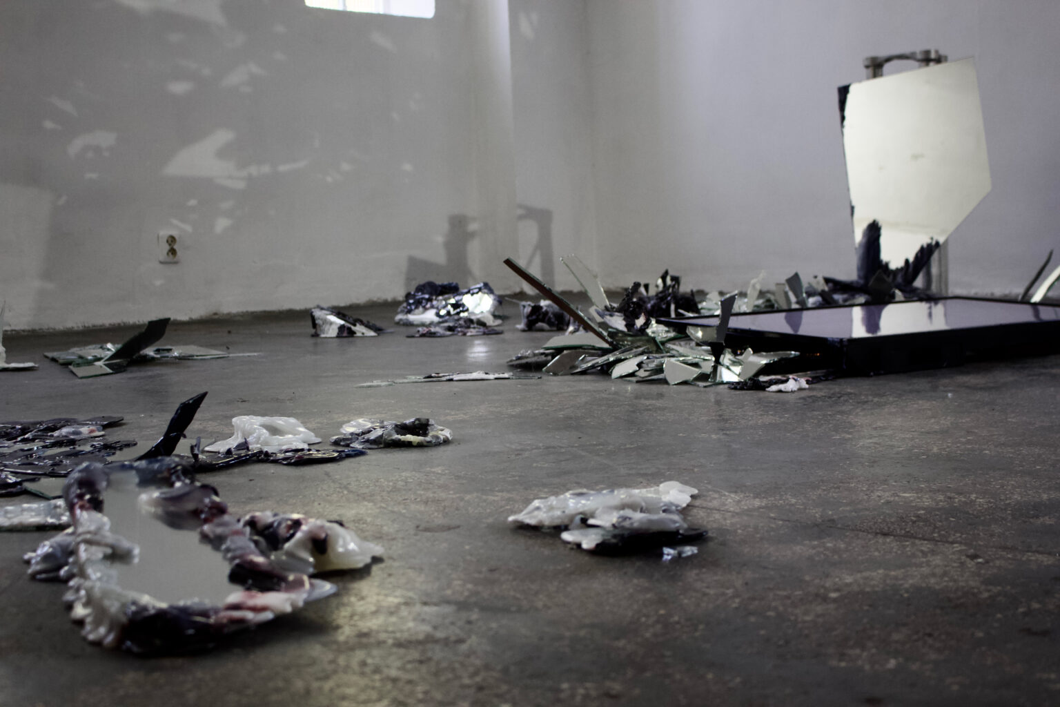 Zofia Mierzejewska: widok wystawy, instalacja. Na podłodze wiele drobnych obiektów z potłuczonego szkła i plastiku, niskie słupy estradowe, położony ekran TV.